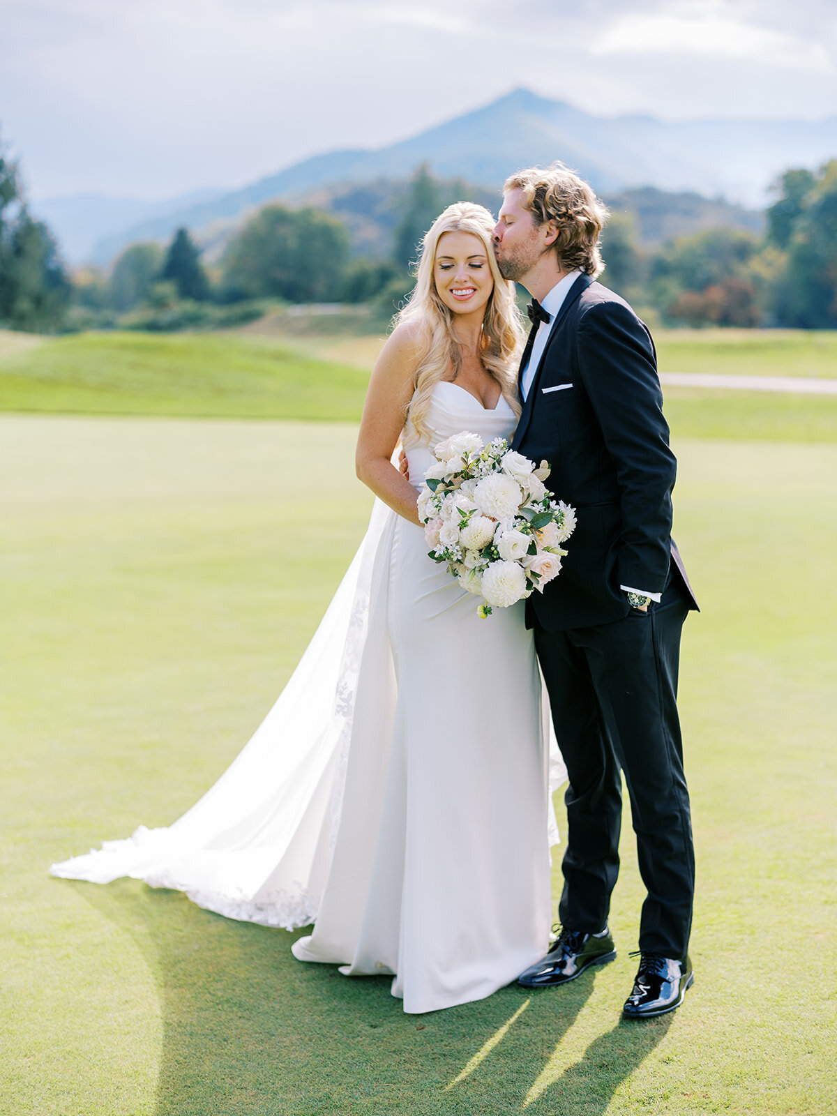 Bride holding bouquet while groom kisses her forehead on the golf course at The Waynesville Inn & Golf Club in Waynesville, North Carolina.
