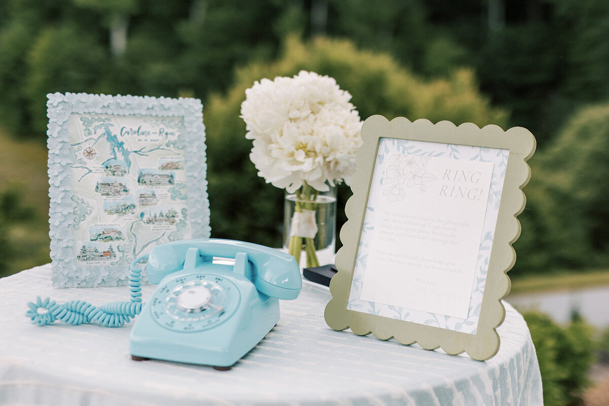 Audio guestbook display featuring a blue rotary phone, watercolor venue map, and white floral arrangement on linen-covered table.
