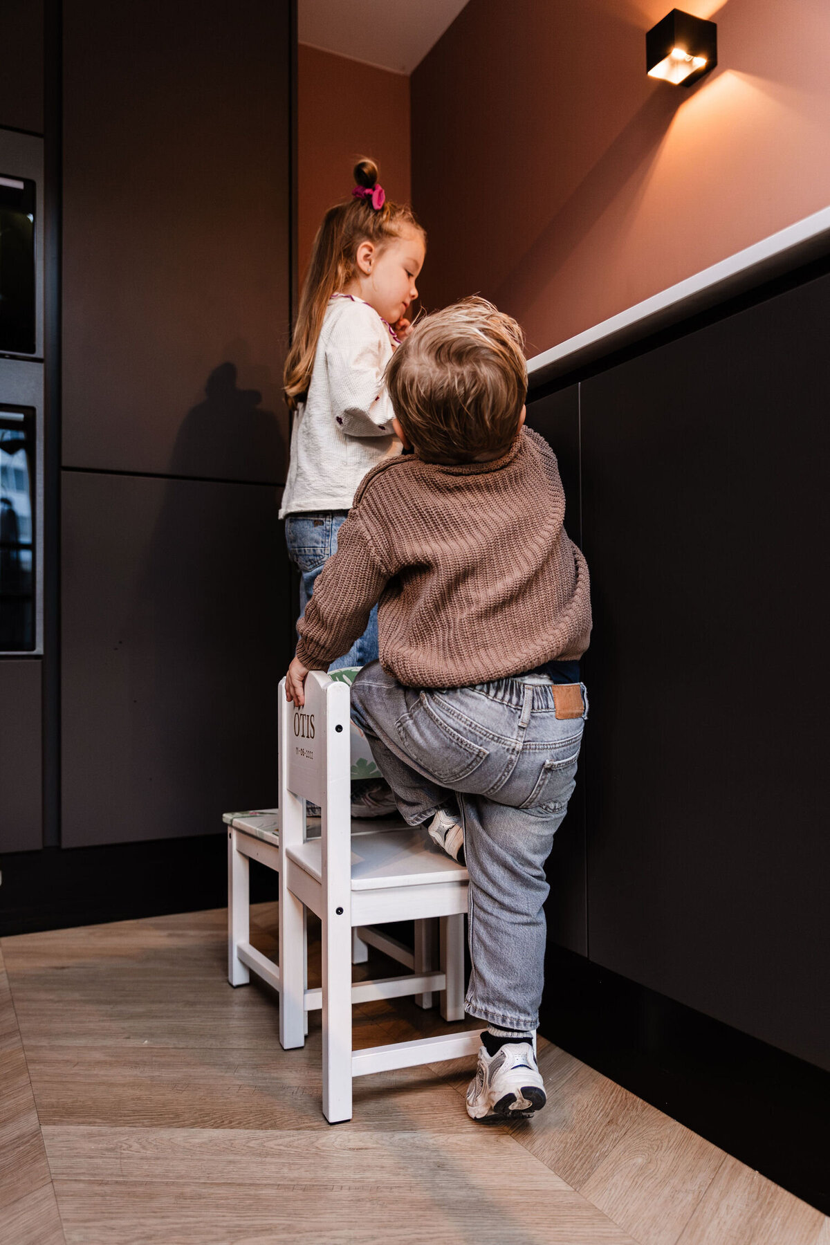 Meisje staat aan aanrecht op klein stoeltje en broertje schuift klein stoeltje bij om naast haar te gaan staan