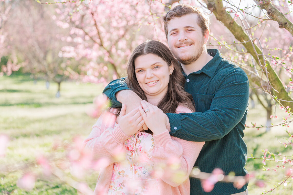A happy engaged couple dresses in bright pastels surrounded by bright pink cherry blossoms in the North Carolina countryside enjoying their destination wedding photos