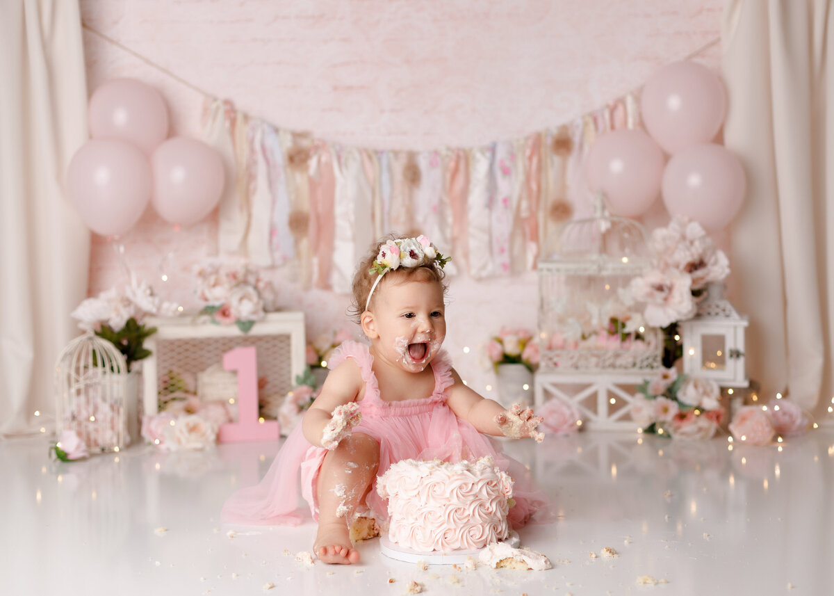 Shabby chic cake smash in West Palm Beach in Wellington photography studio. Baby girl is wearing a pink tutu dress behind a soft pink cake with frosting on her face, hands and feet. The background is a soft pink with cream curtains on either side surrounded by a floral banner and floral accents throughout.