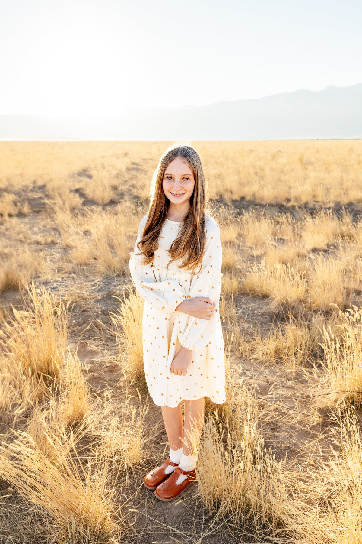 A young girl with long blonde hair wearing a white dress with small pumpkin prints stands in a sunlit field in Eagle Mountain, Utah.