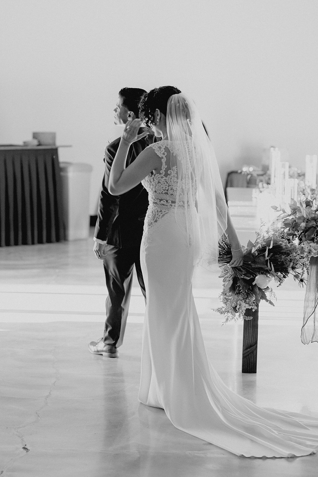 Black and white photo of the bride and groom from behind, walking together with the bride holding a large bouquet, capturing a quiet, intimate moment.