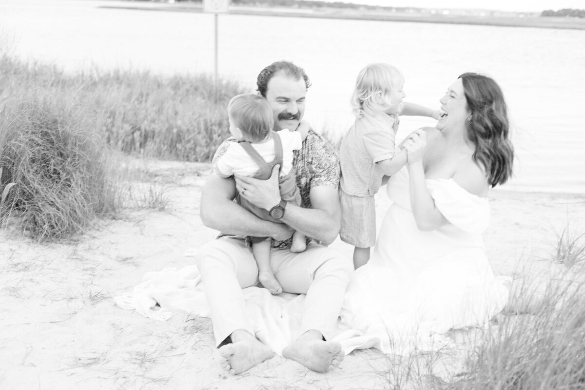 Black and white Virginia Beach Maternity Photography portrait of a young family sitting on a blanket on the beach playing together.