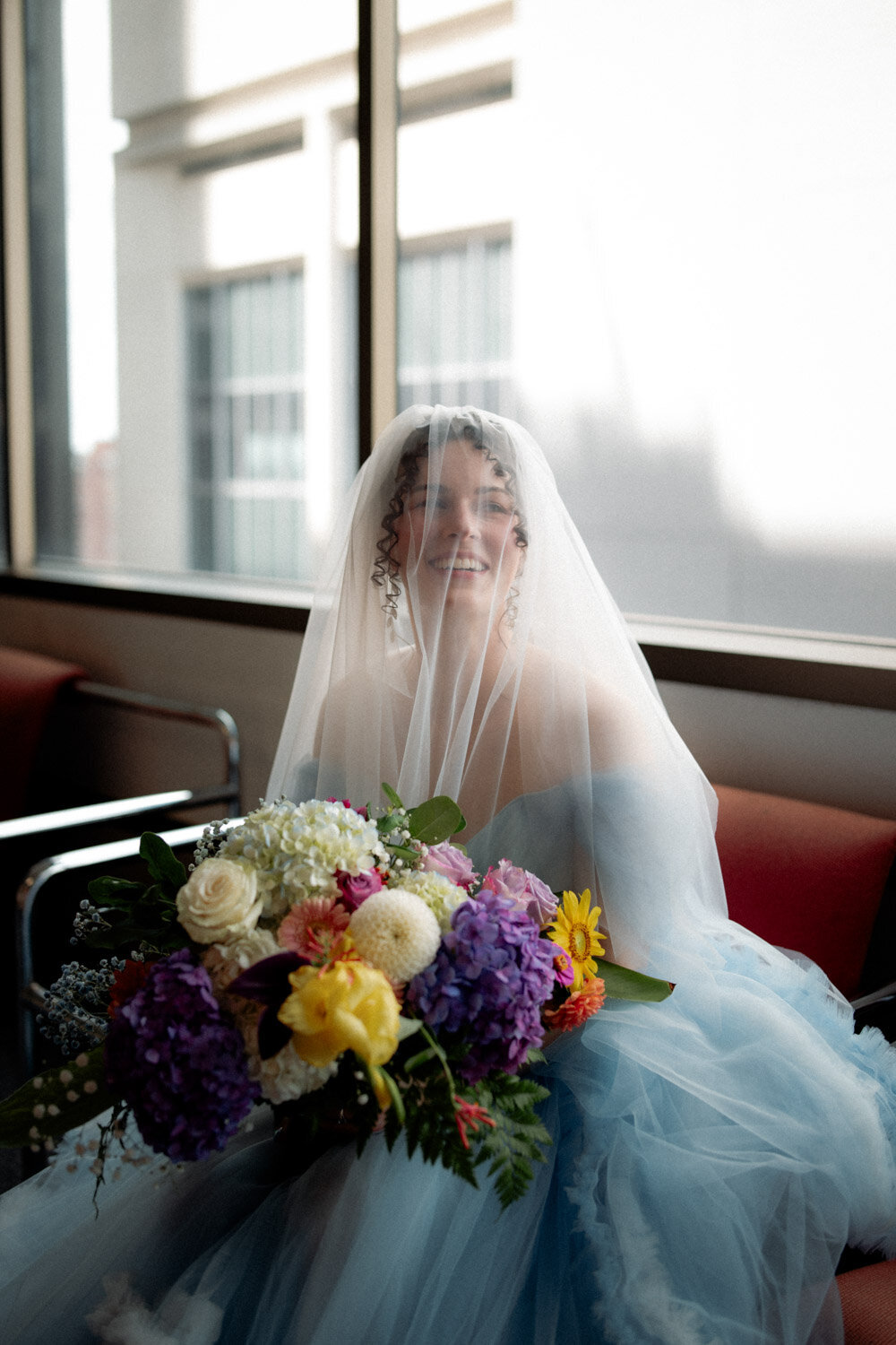 Bride waits outside courthouse with blue dress and veil covering face