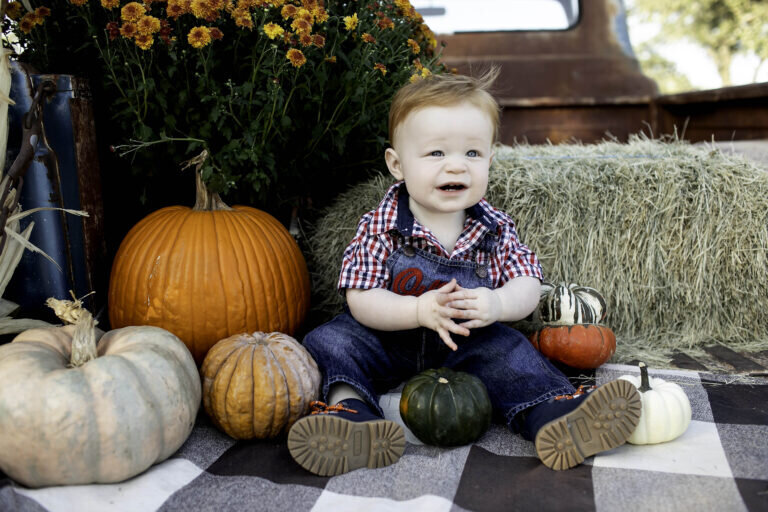 First birthday photoshoot with a baby boy standing beside a pile of pumpkins in the patch, looking curious and happy