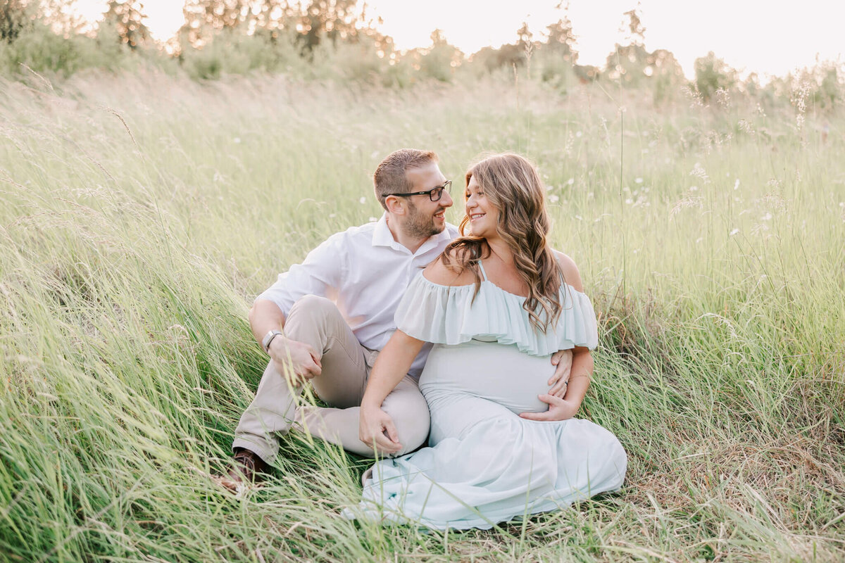 portrait of husband and wife in oregon field smiliing with hands on belly. She is wearing a green maternity dress.