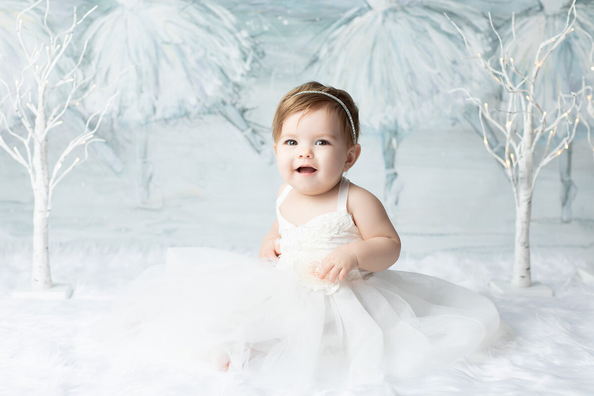 Dallas milestone photography featuring a baby girl in a white gown sitting on faux snow, framed by birch trees and ballerina artwork in soft wintry tones.