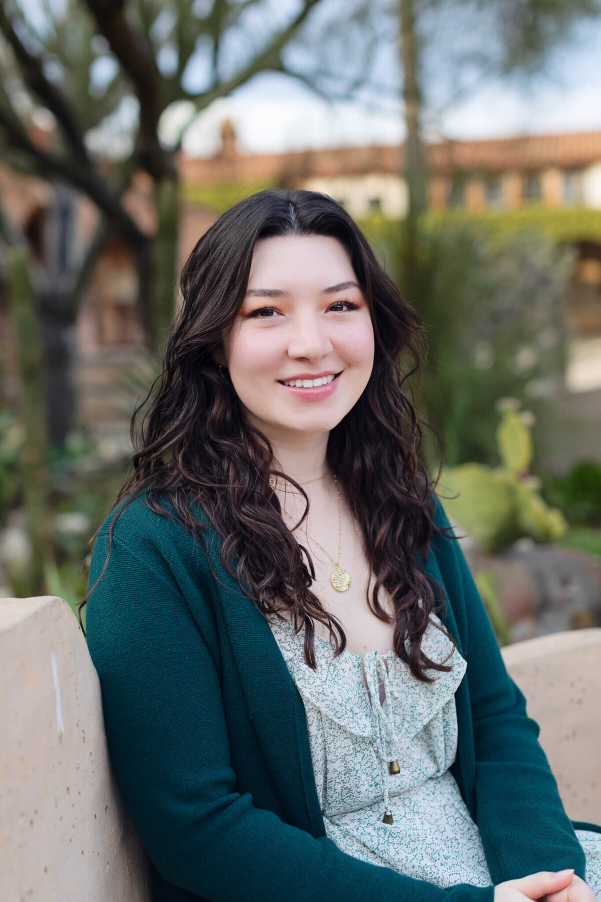 A close-up of a beautiful young woman with black hair, wearing a green cardigan, looking at the camera and smiling in a courtyard of greenery.