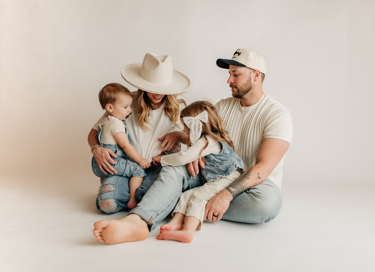 Family of 4 sitting on the ground in a white studio setting, all looking at each other laughing and playing