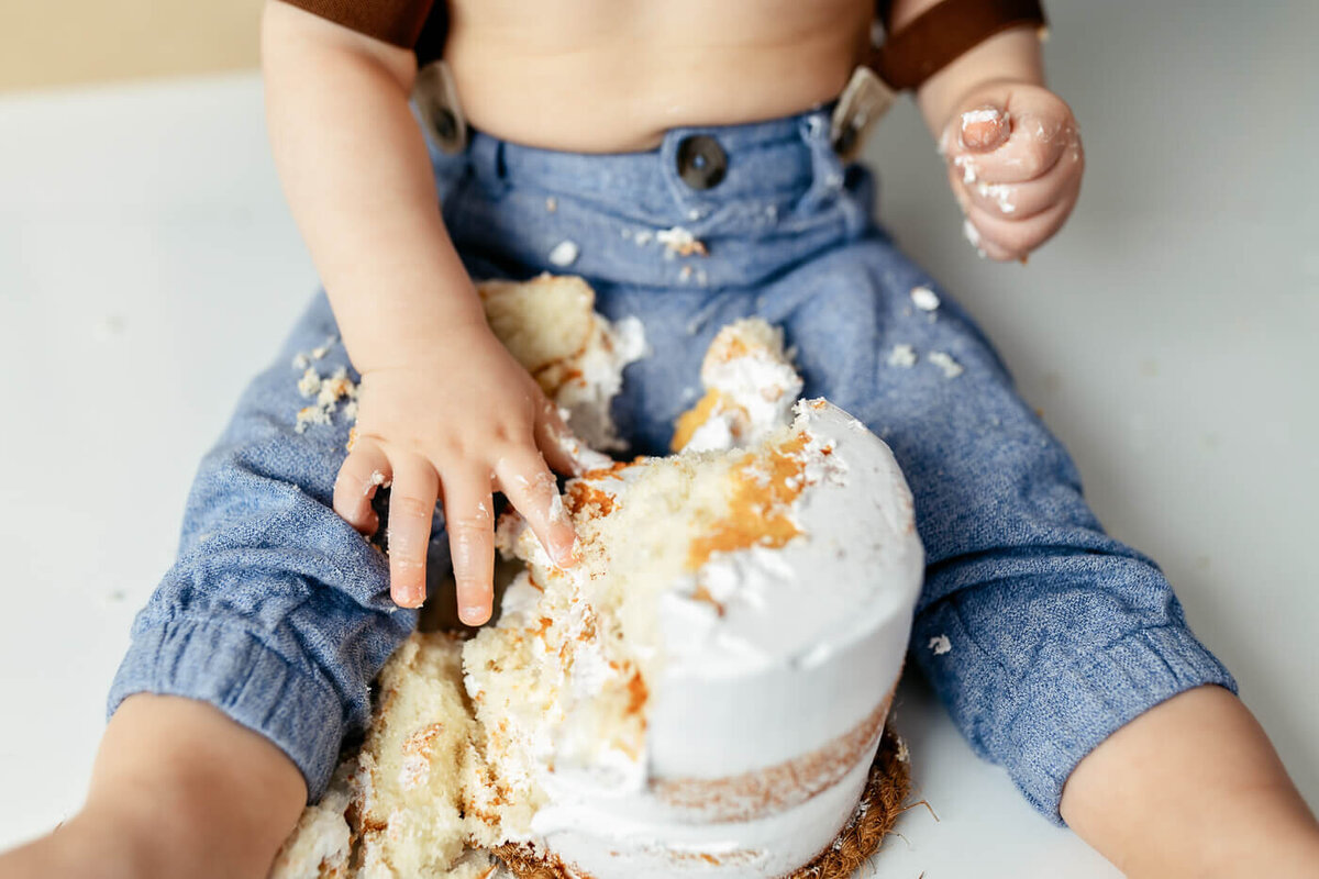A baby sitting on the floor explores a partially eaten cake with both hands. Crumbs and frosting cover the baby's denim pants, showing they are enjoying a playful and messy moment.
