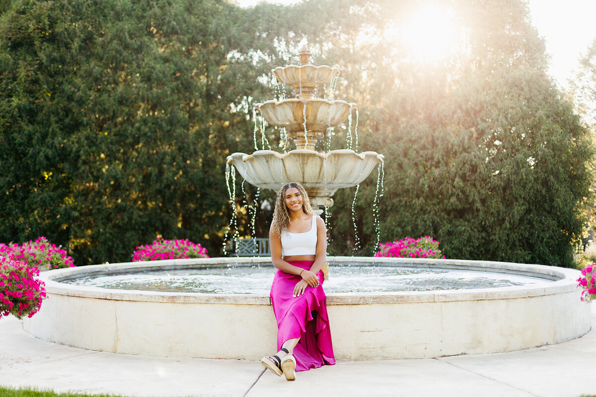 Senior girl in a white top and pink skirt sitting in front of a fountain