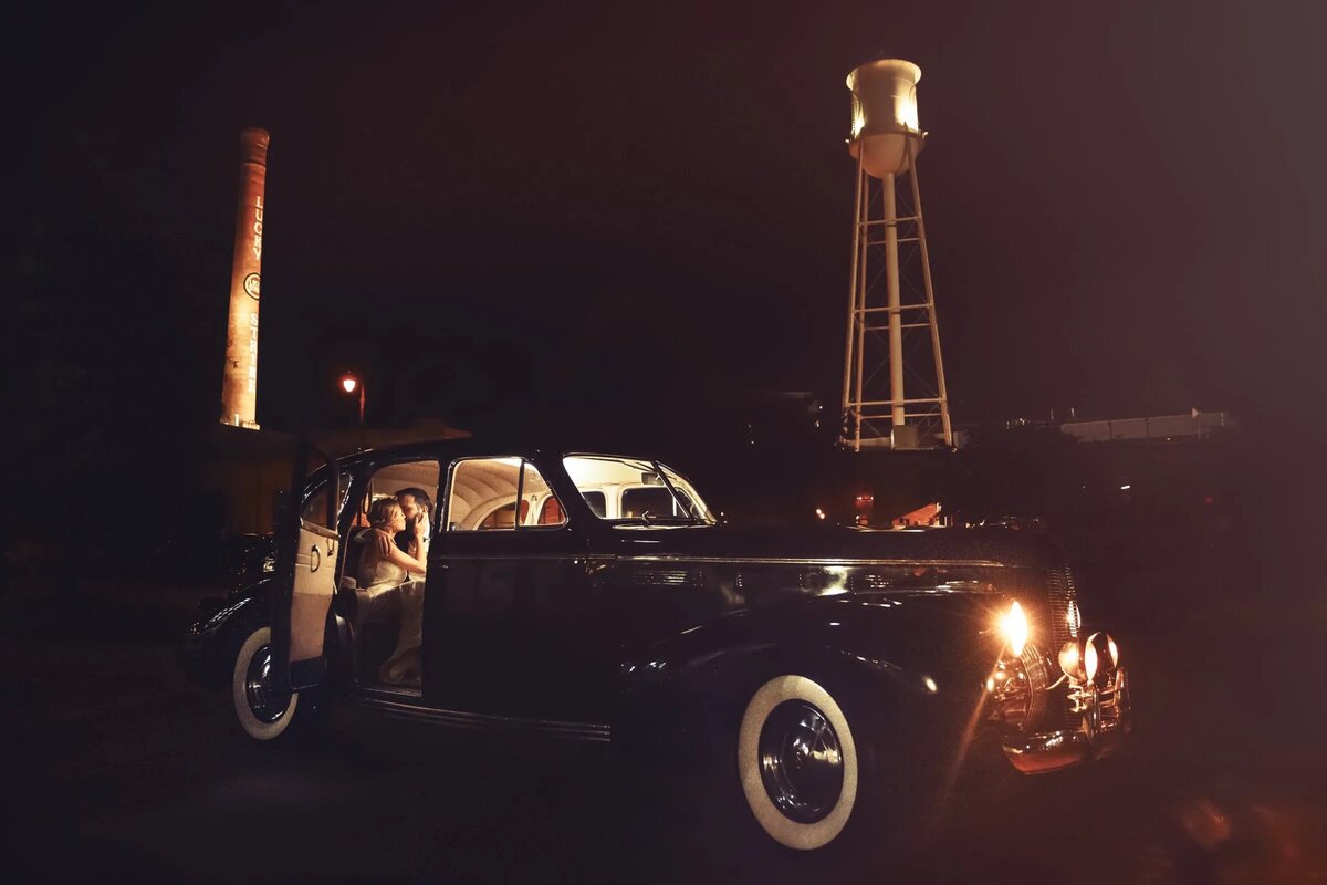 Vintage car parked at night in front of a lit water tower, giving off a classic and timeless vibe with a bride and groom sitting in the back