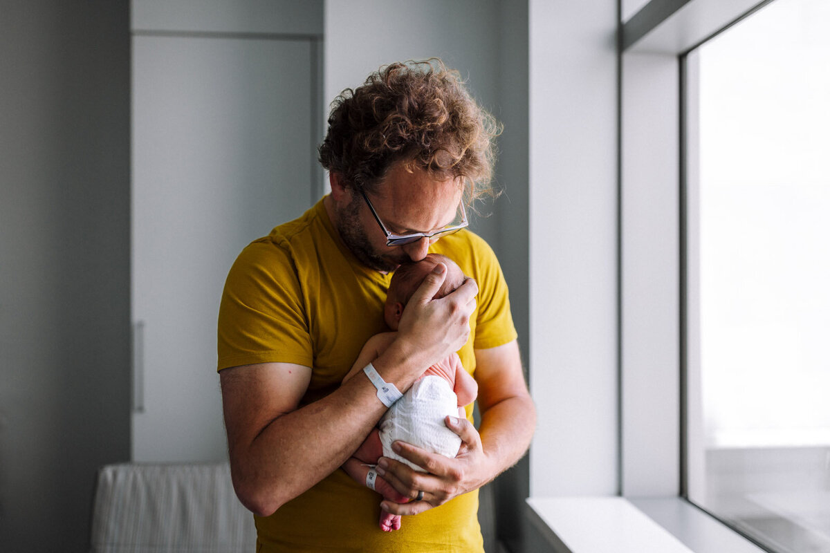 dad in golden shirt holding newborn in diaper to his chest and kissing his forehead