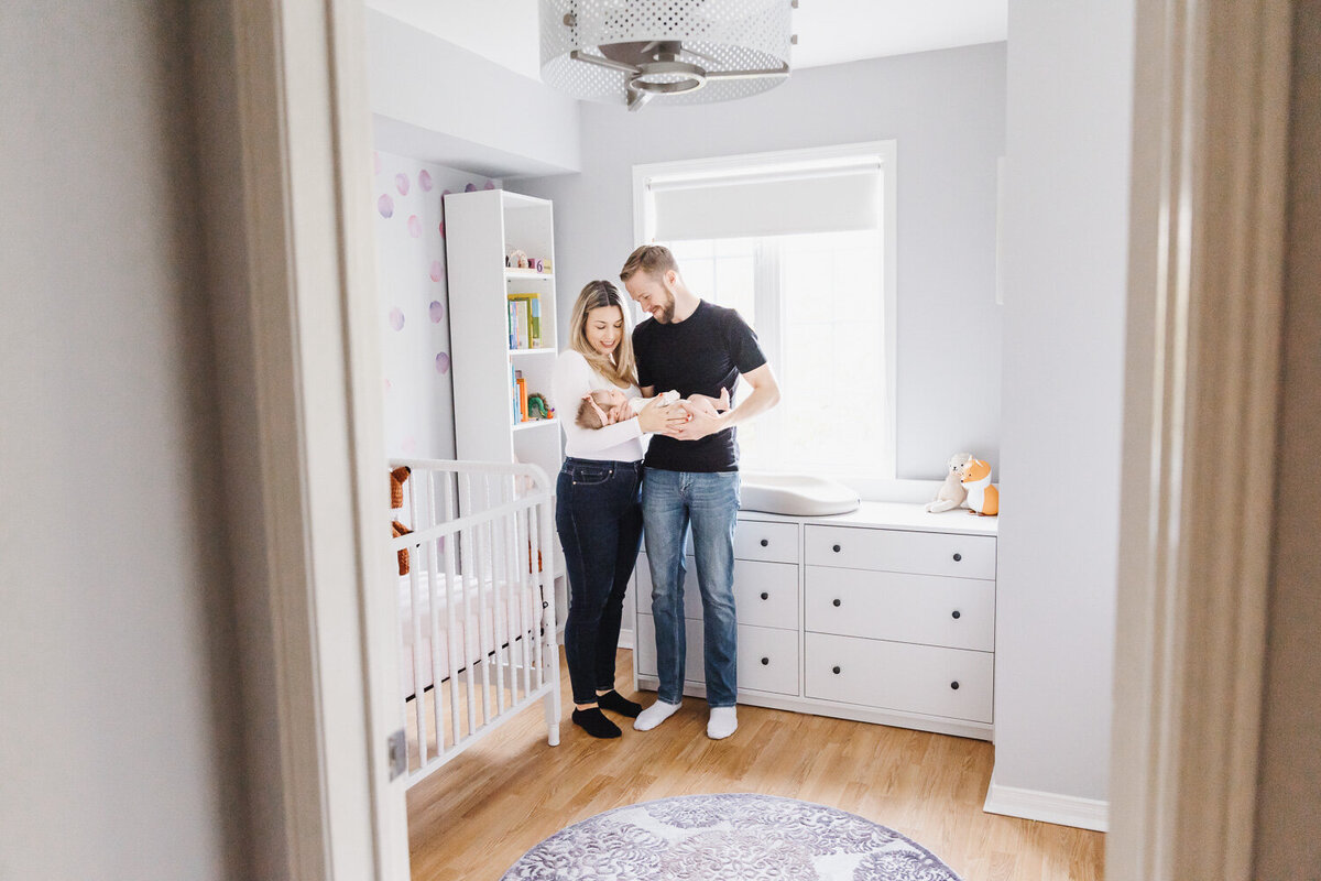Parents in nursery standing by window holding newborn daughter and looking down at her smiling
