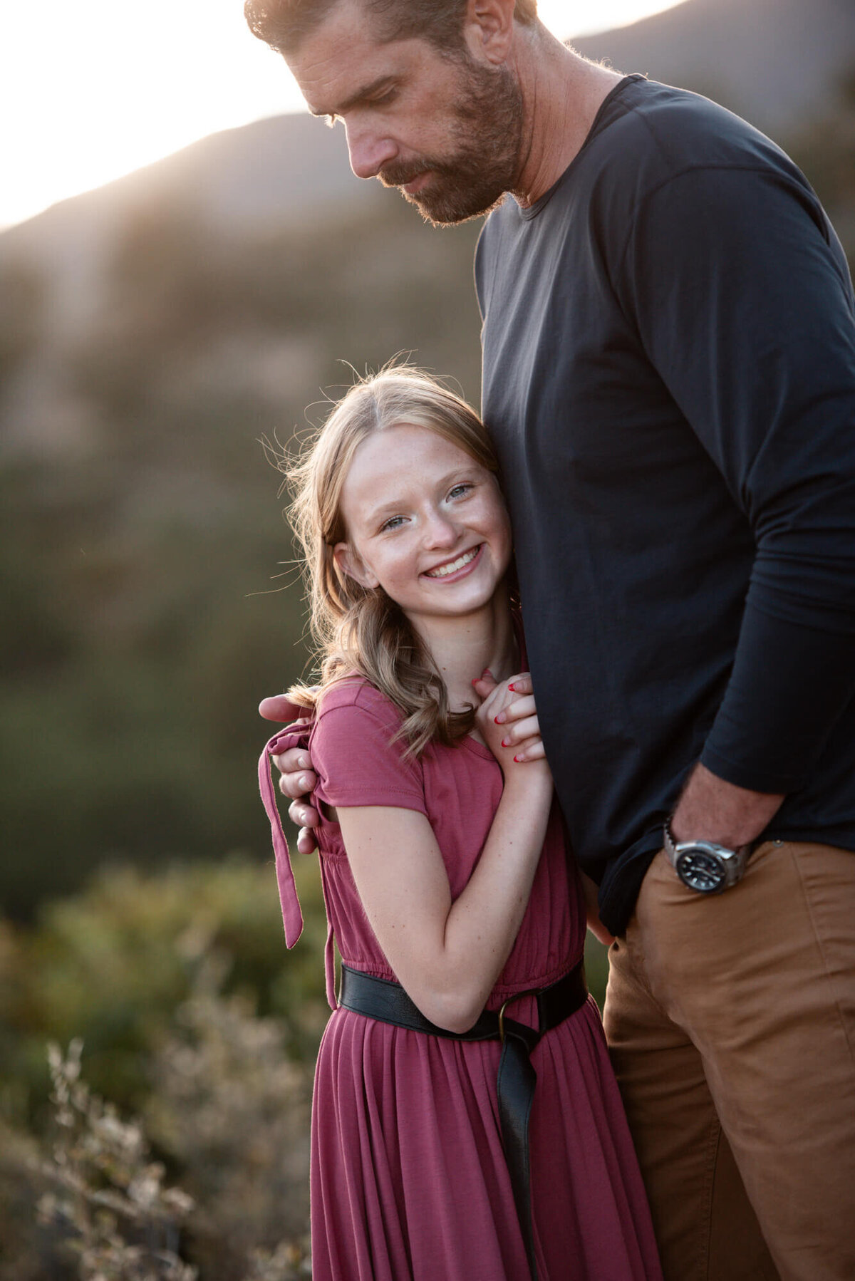 A young girl in a pink dress hugs onto her father as they stand in a field of tall grass