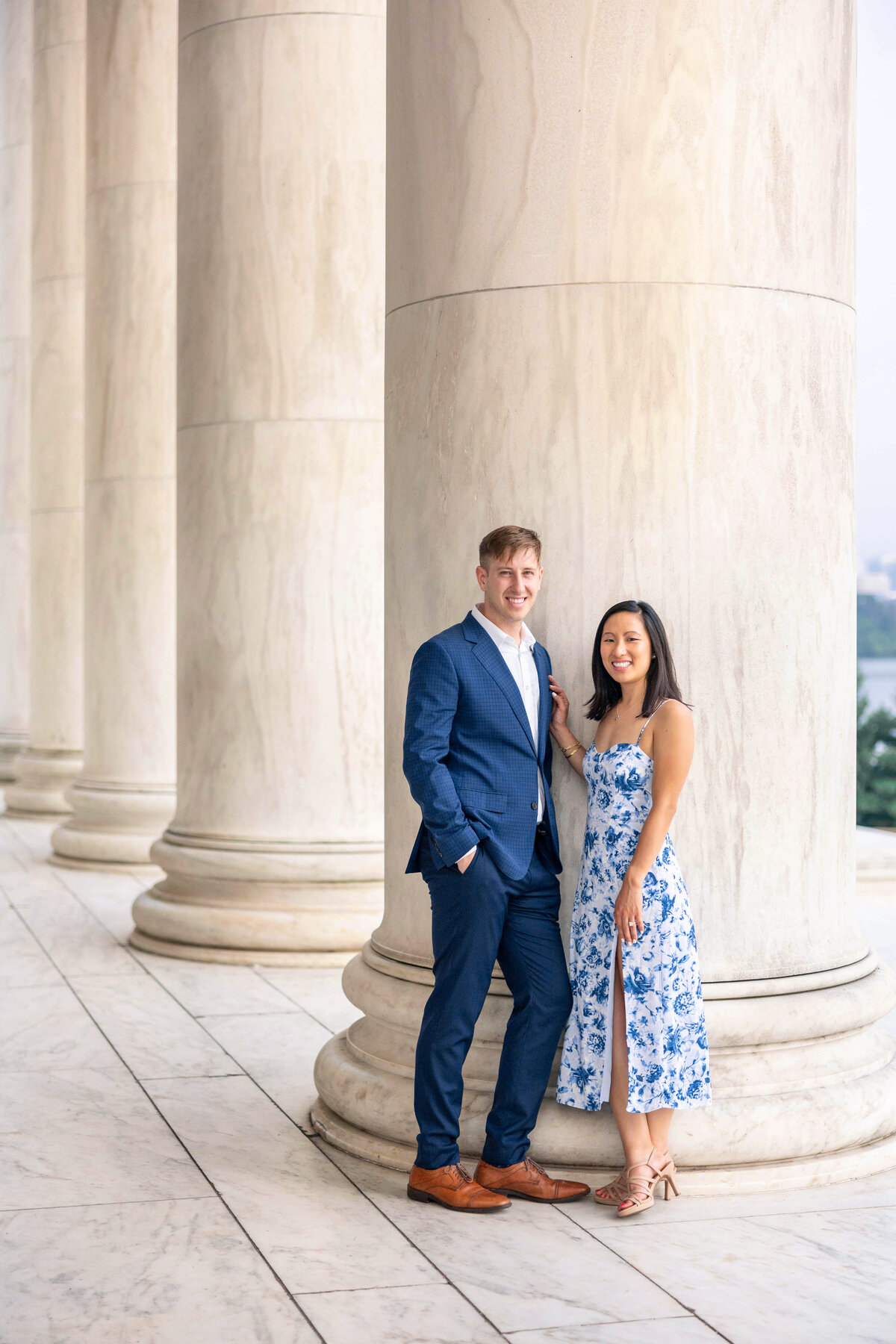 A man in a blue suit and a woman in a blue floral dress stand smiling next to a large marble column. The setting is outdoors with classical architecture in the background.