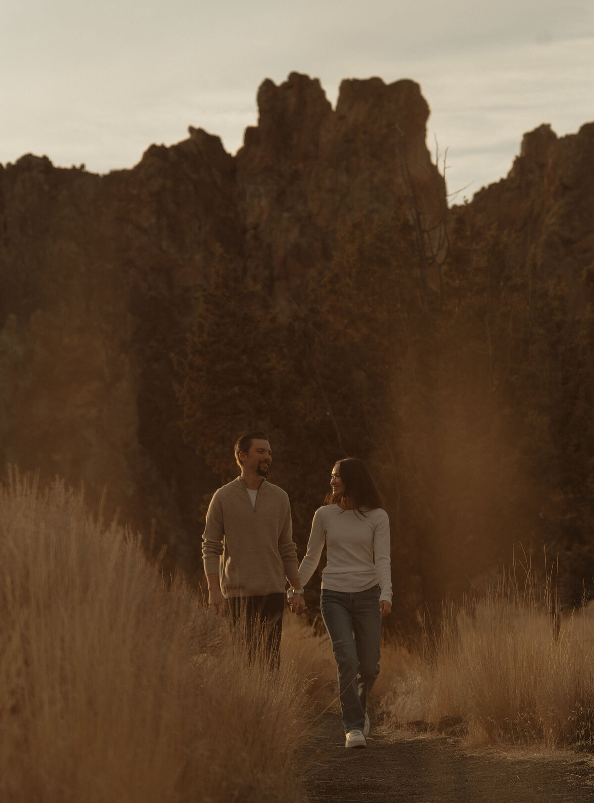 engaged couple holds hands while looking at each other and walking the trails at Smith Rock