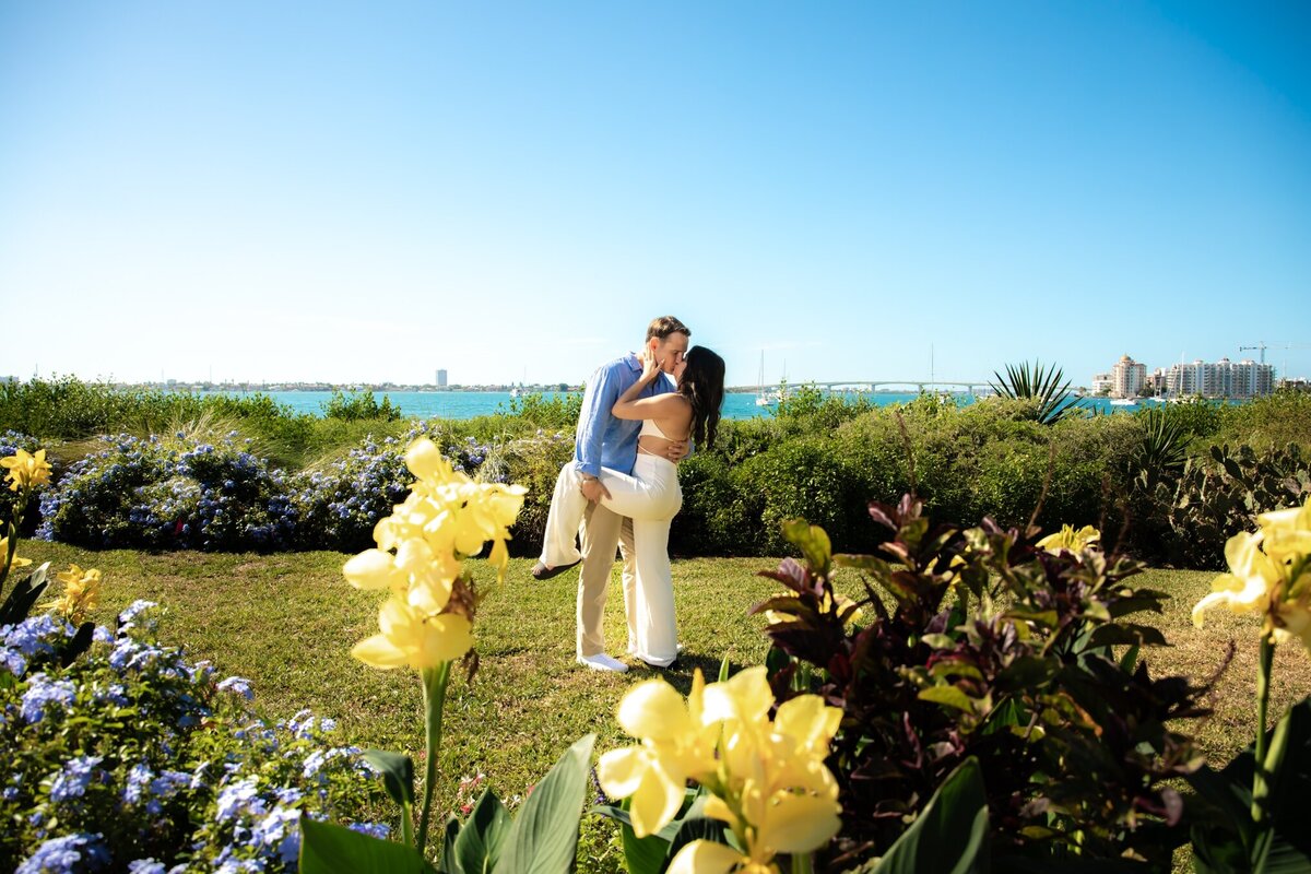 Engaged couple kissing at Selby Gardens with flowers in the foreground and Sarasota Bay in the background
