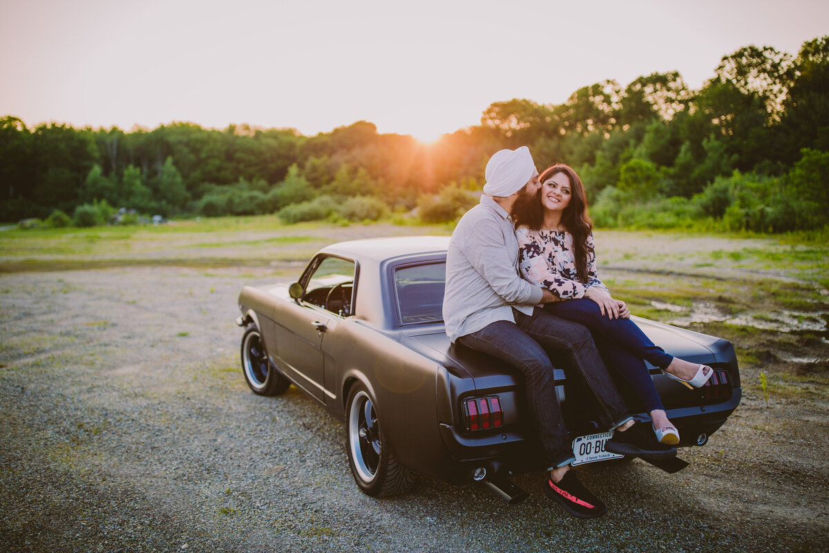 An engaged sikh couple sitting on top of the trunk of an old vintage ford mustang
