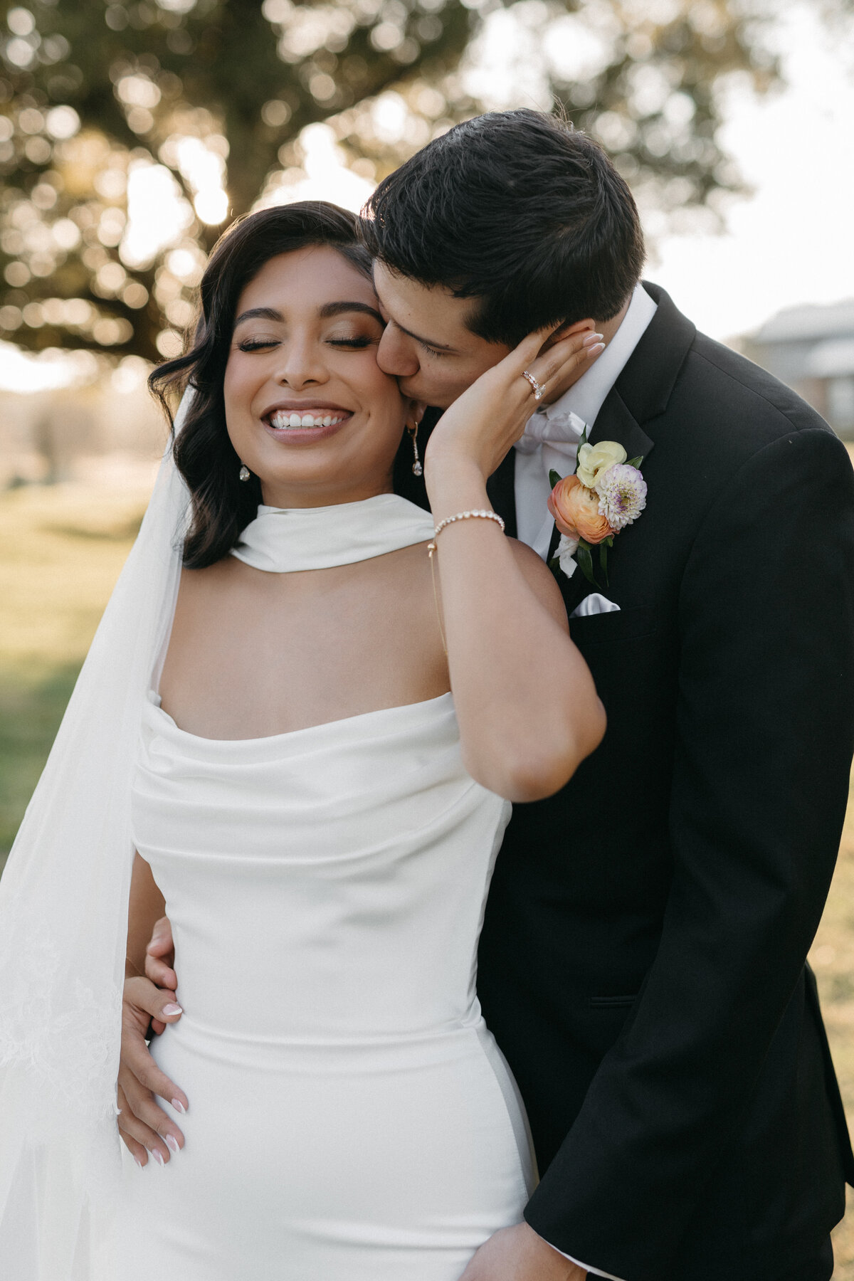 Romantic outdoor wedding portrait showcasing the groom's peach and ivory ranunculus boutonniere beside the bride in an elegant white gown, captured during golden hour for soft, natural floral-focused wedding inspiration.
