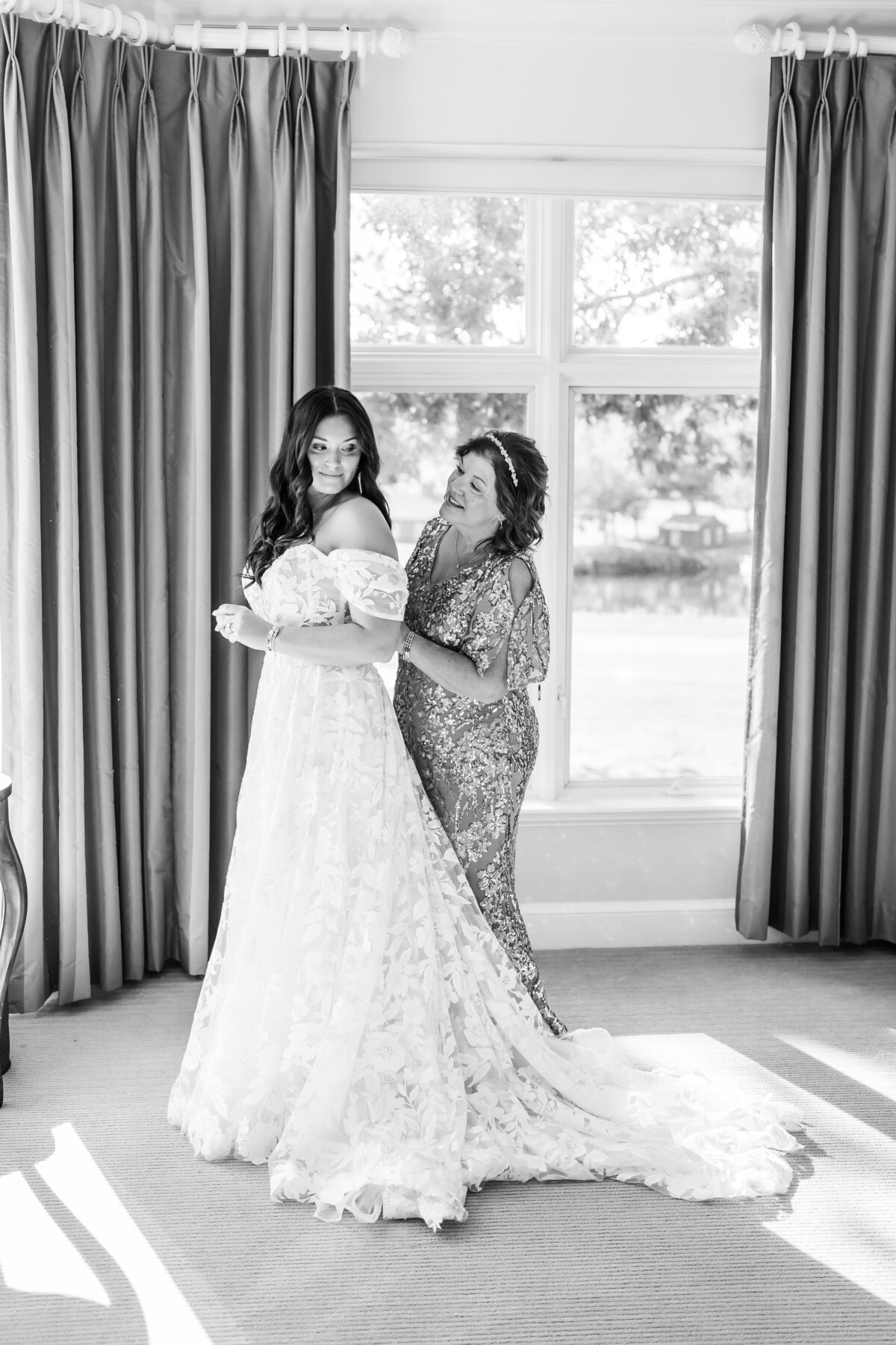 Timeless black and white wedding photography capturing a heartfelt moment between a bride and her mother. The bride, in a romantic off-the-shoulder lace gown, shares a joyful, emotional moment as her mother helps her get ready. Natural light fills the elegant bridal suite, highlighting the love and anticipation of this special day. Captured by Moll – Boston wedding photographer specializing in authentic and emotive storytelling.