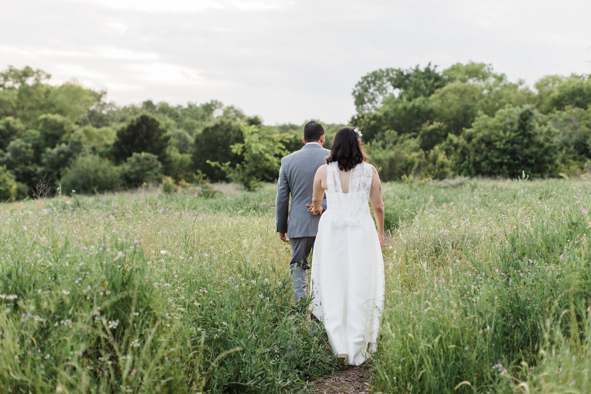 A shot of a bride and groom walking away from the camera in a field of tall grass after their wedding ceremony at the Trinity River Audubon Center in Dallas, Texas. The bride is on the right and is wearing a sleeveless, intricate, white dress. The groom is in front and to the left of her and is wearing a grey suit.