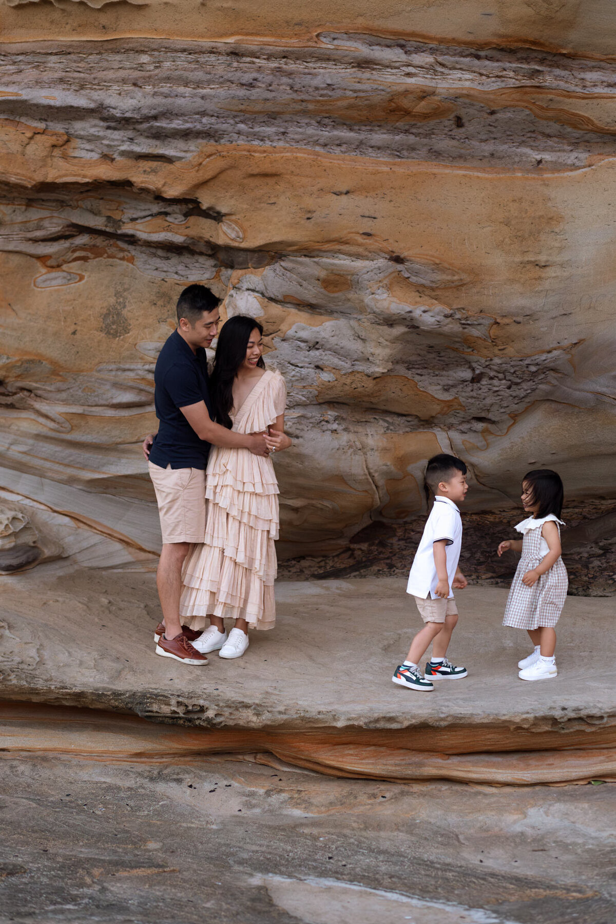 Family of four sharing a joyful moment by a textured sandstone wall, with parents embracing and children laughing together during a beachside family photoshoot.