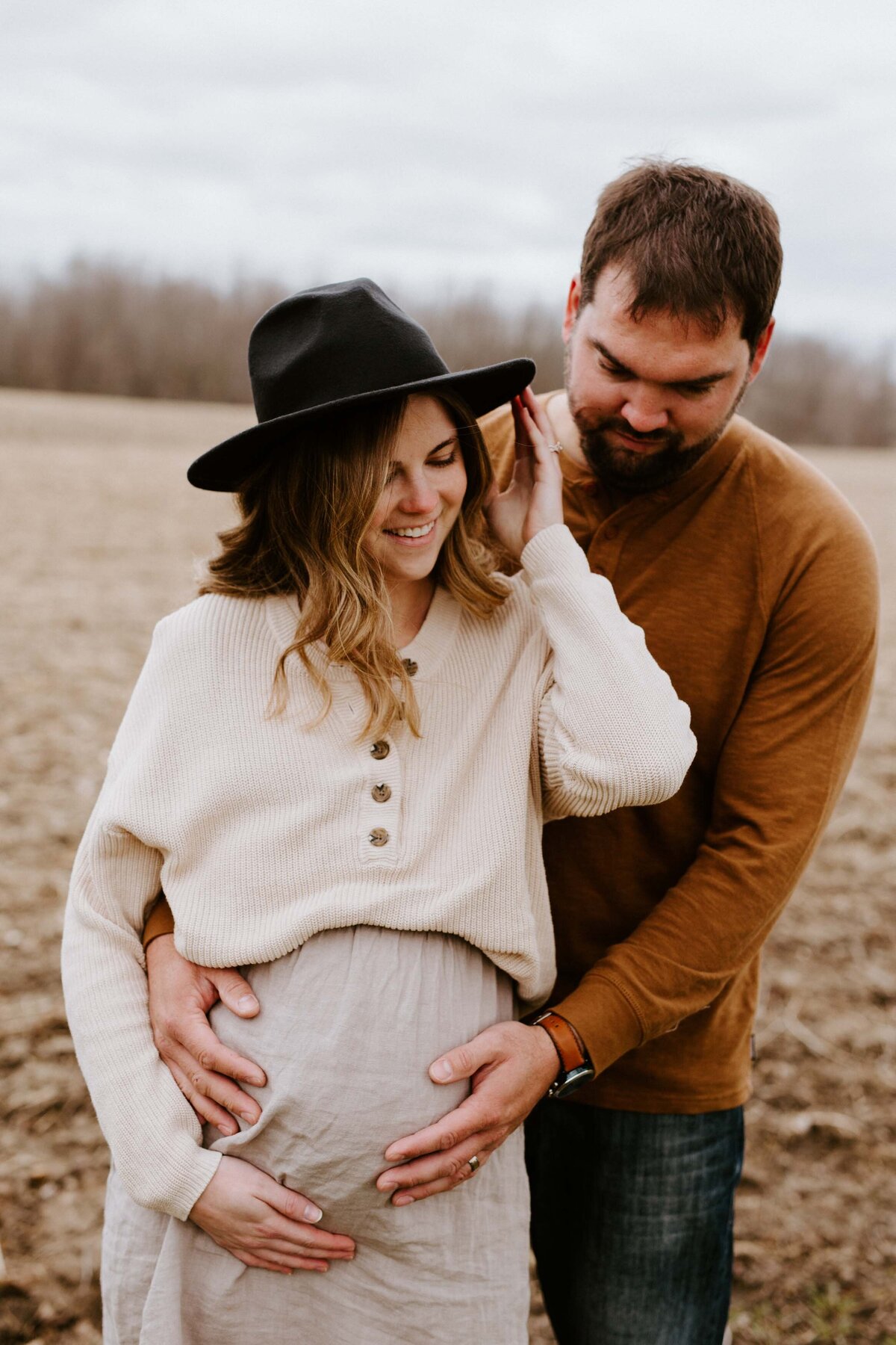 Expectant mom and Dad in Exeter, Ontario farmers field for maternity photo session. Mom is facing the camera, dad is behind mom holding her bump. Mom has one hand on her belly and the other is touching her hat. Both mom and dad are smiling at her belly.