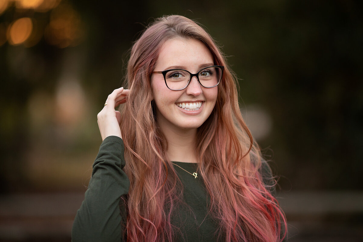 A smiling young woman with pink highlights in her hair, wearing glasses and a green top, captured outdoors in a family photography session.