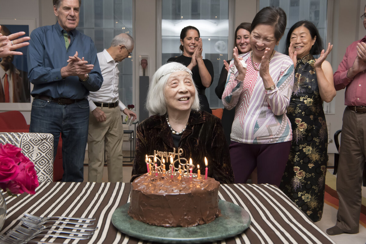 A family cheering while their grandparent sits at a table in front of a birthday cake 
