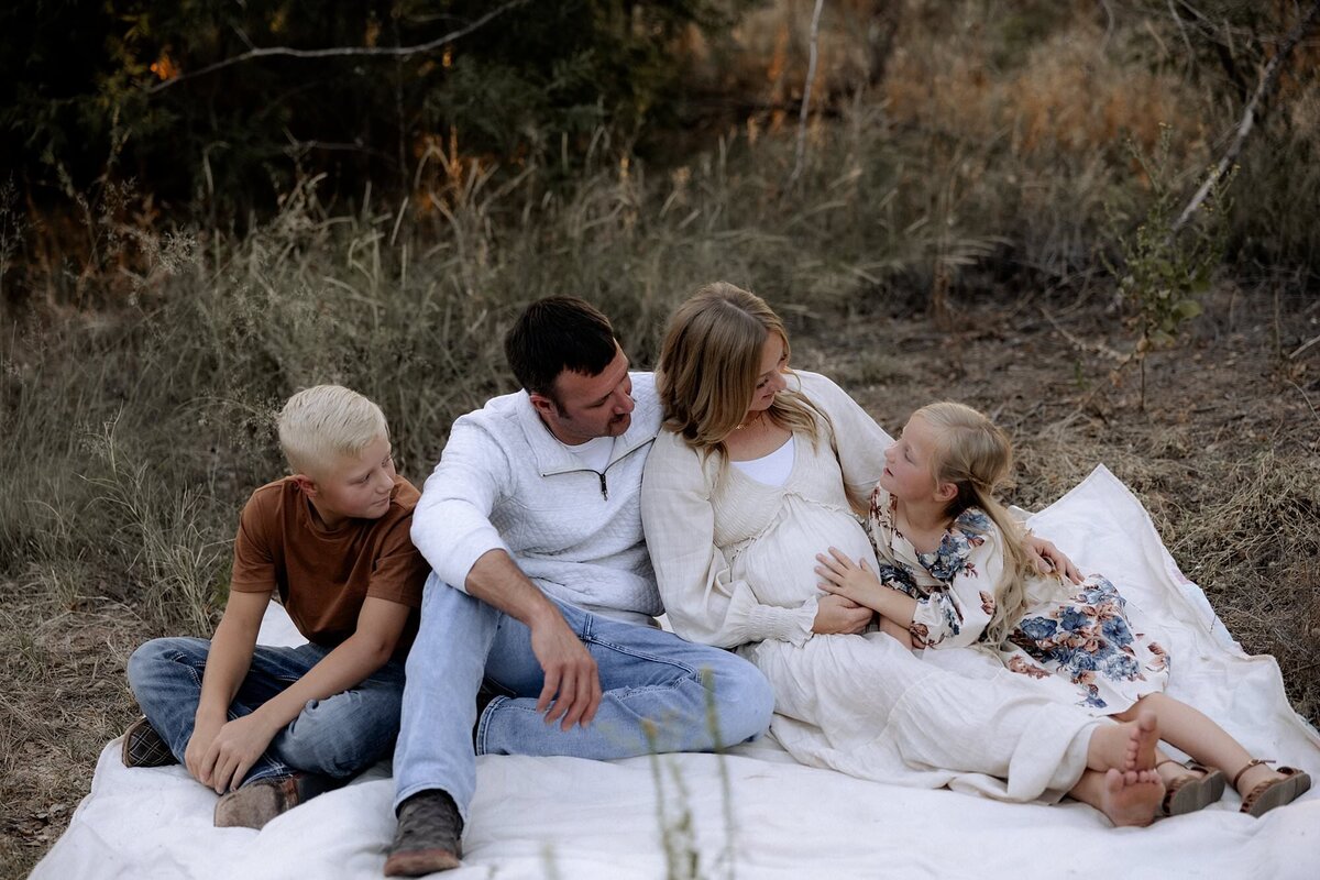 maternity photo of family of four sitting on blanket on grass, sister touching mama's pregnant belly