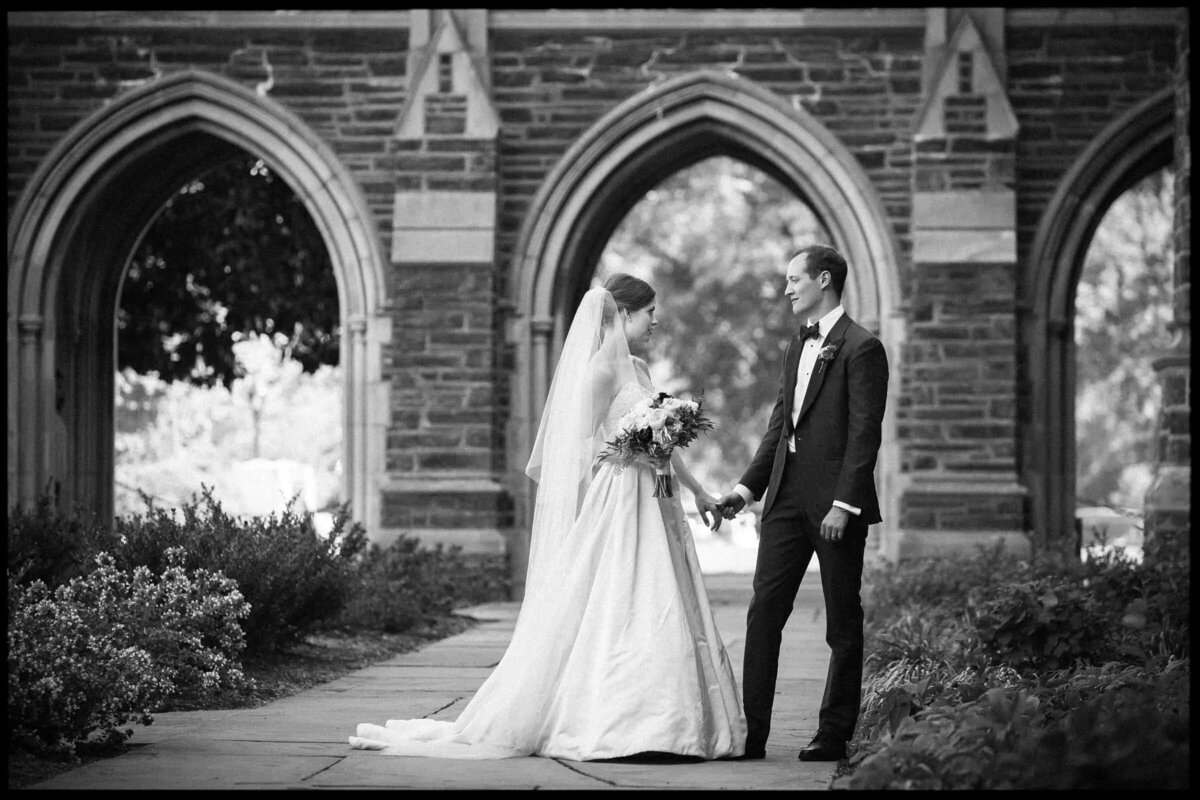Bride and groom standing hand in hand, exchanging a loving look in an outdoor garden with beautiful arches