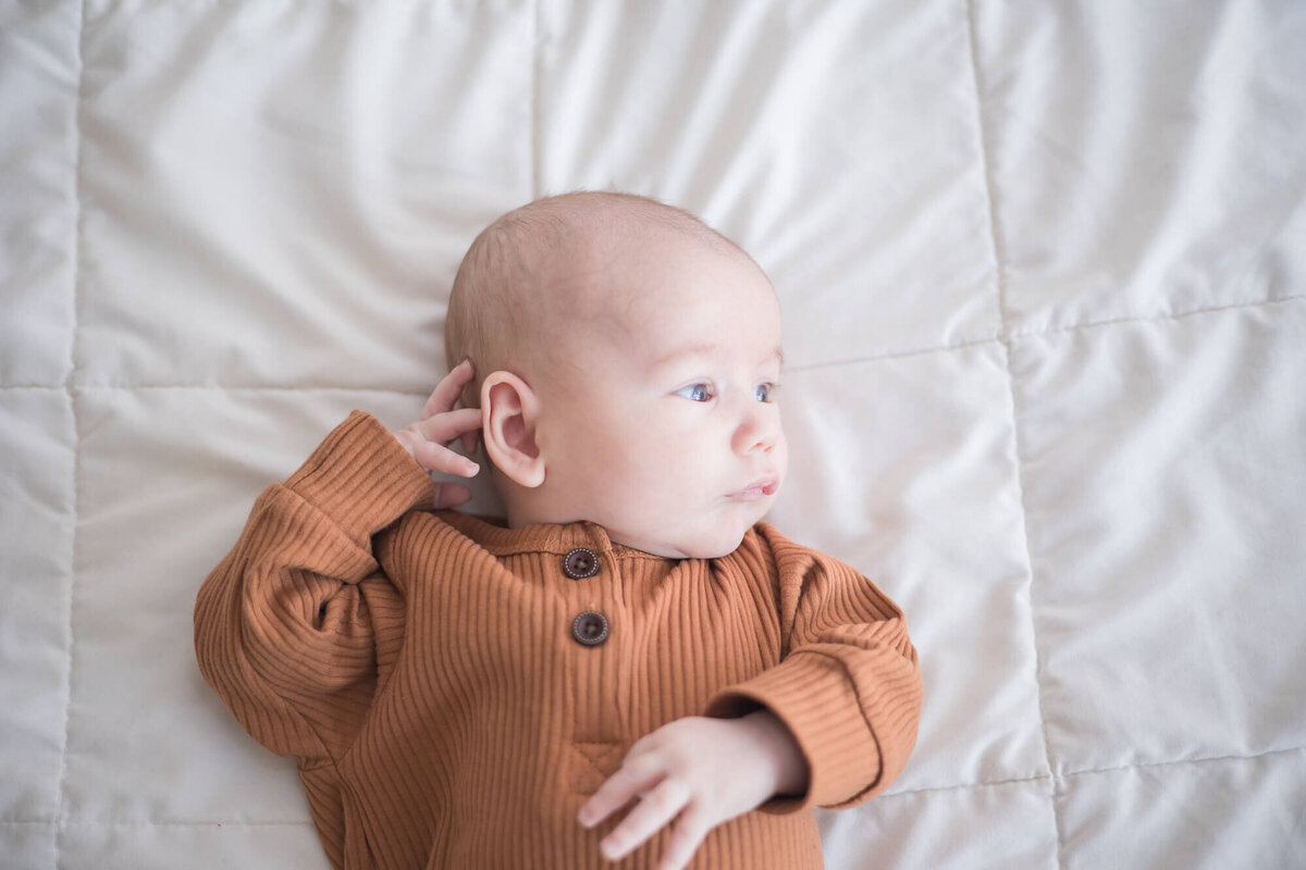 newborn infant baby boy laying on a white bed looking to the side while touching his ear.