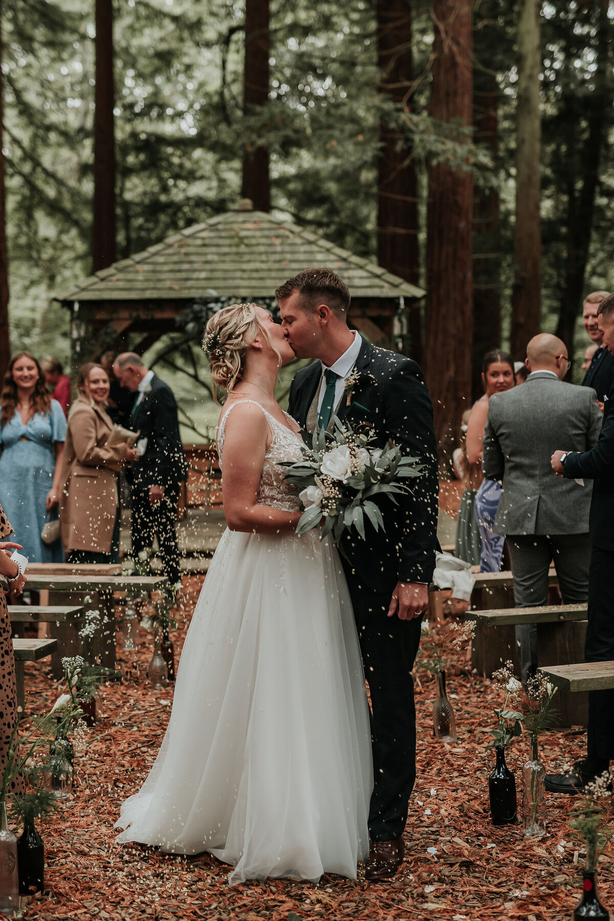 Bride & Groom kiss amongst confetti in the aisle at their woodland ceremony at Two Woods Estate