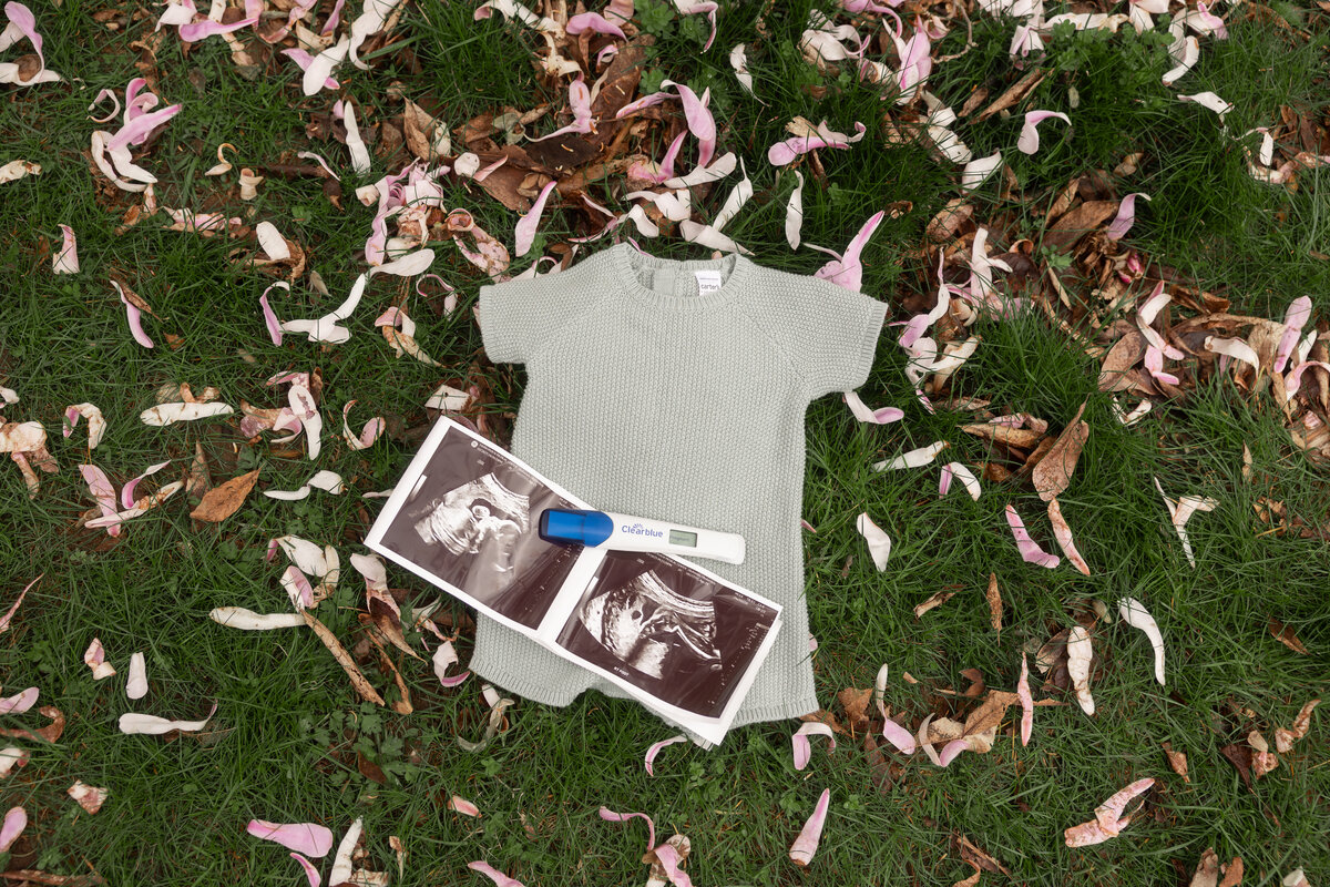 Flat lay of a pregnancy announcement featuring a small knitted baby outfit, ultrasound images, and a Clearblue positive pregnancy test, arranged on green grass with scattered pink flower petals.