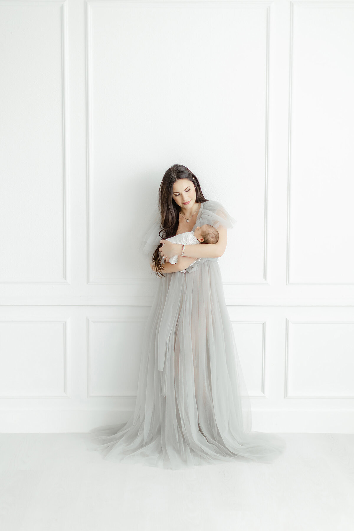 An ethereal portrait of a mother standing in a Dallas photography studio dressed in a light grey tulle gown that's drapped on the floor as she holder her newborn baby for her new family portraits.