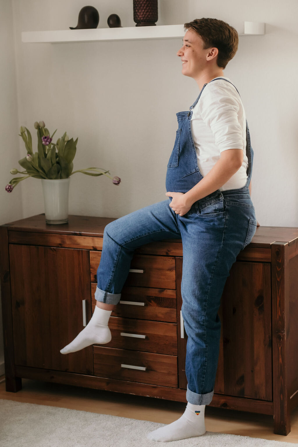 A pregnant woman sits loosely on a sideboard, smiling while holding her baby bump.