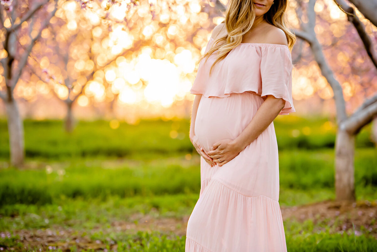 Prengant woman close up of her belly in a pink dress with the golden sunlight behind her for her Burlington, Ontario maternity photography