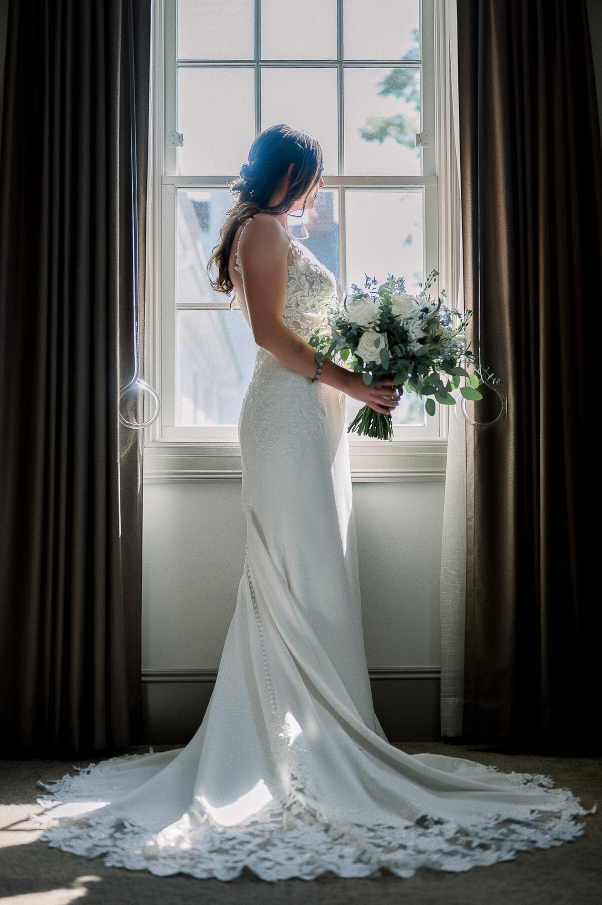 a bride standing in front of a window in her wedding dress while holding a bouquet