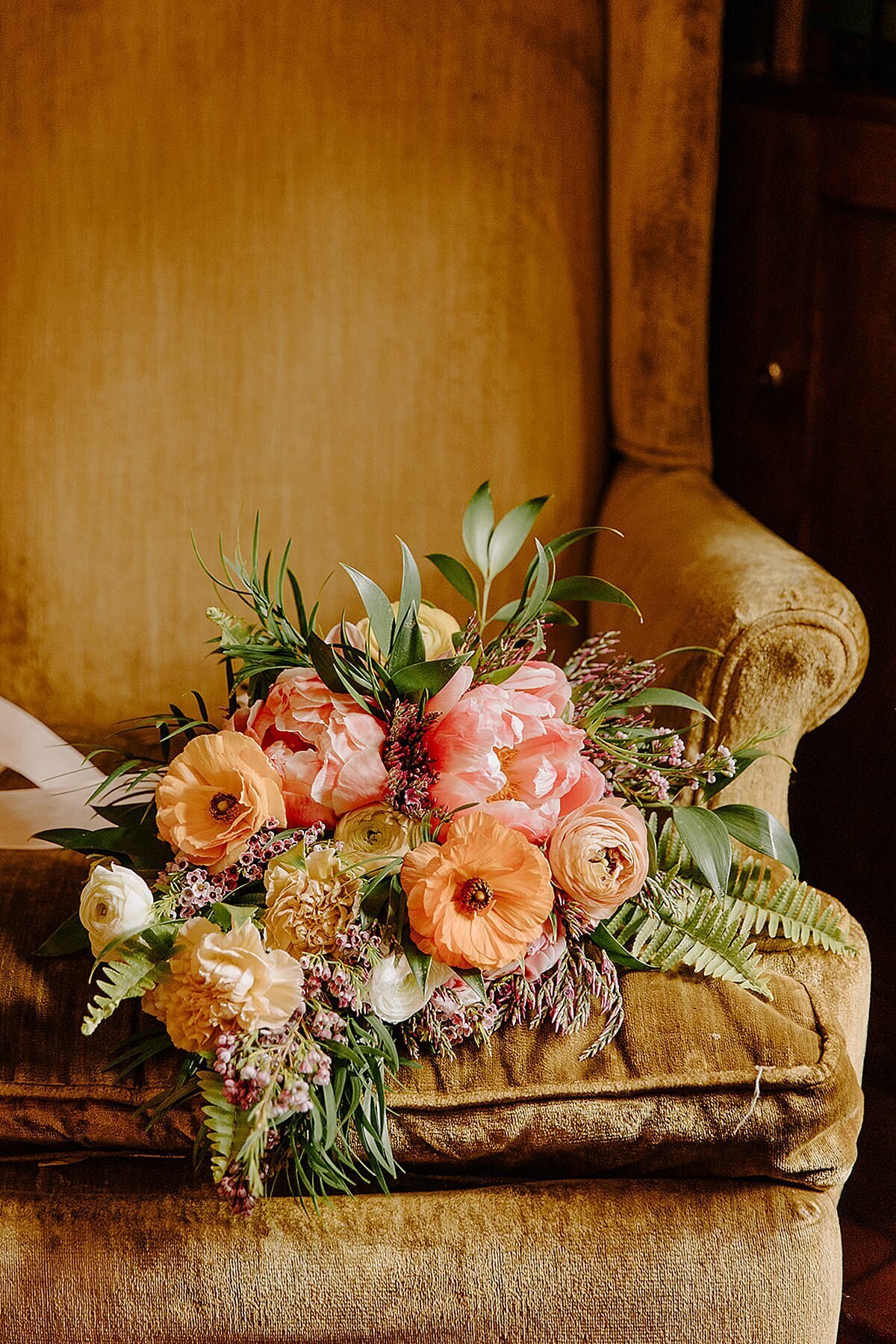 Mustard gold velvet wingback chair with a large bridal bouquet of orange ranunculus, pink peonies, fern, eucalyptus, ivory ranunculus, light brown carnations purple wax flower and greenery wrapped in ivory satin ribbon.