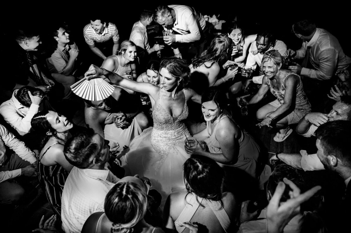 An overhead black and white shot of a bride in a beaded gown dancing joyfully surrounded by guests at a lively wedding reception.