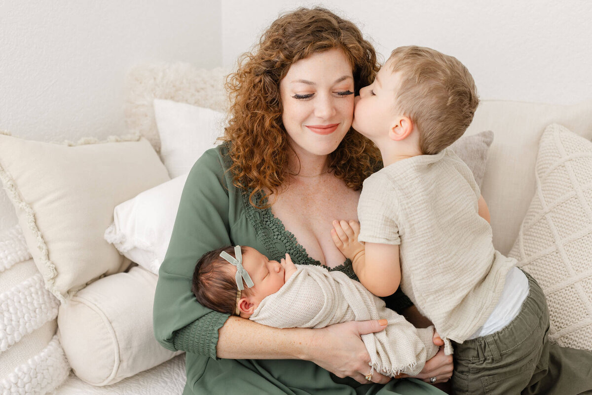 Mom is sitting on a bed while holding newborn baby girl. Toddler son is kneeling next to her and reaching up to kiss her on the cheek. Mom has a soft smile on her face and eyes are closed.
