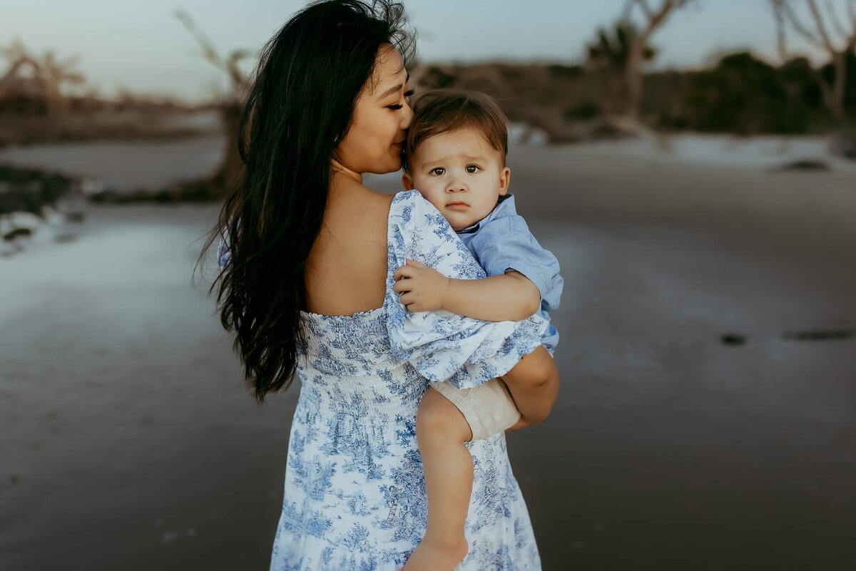 Woman in a blue and white dress holds a toddler wearing a matching dress on a deserted, beach-like area with sparse trees in the background.