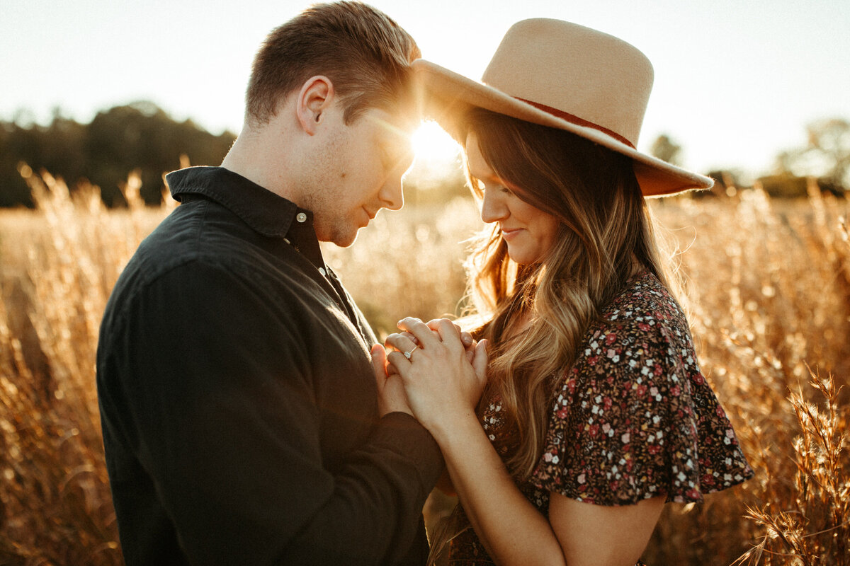 Girl in boho hat and floral dress holding hands with her fiancé in a field as the sun shines between them