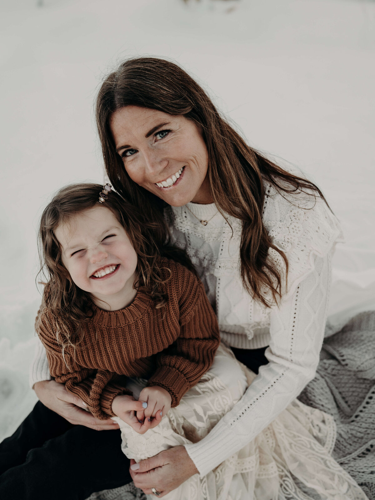 A mother holds her daughter that looks just like her during a family photo session. The daughter is about three years old and is wearing a brown oversized sweater while the beautiful mom wears a white sweater with a snowy background..