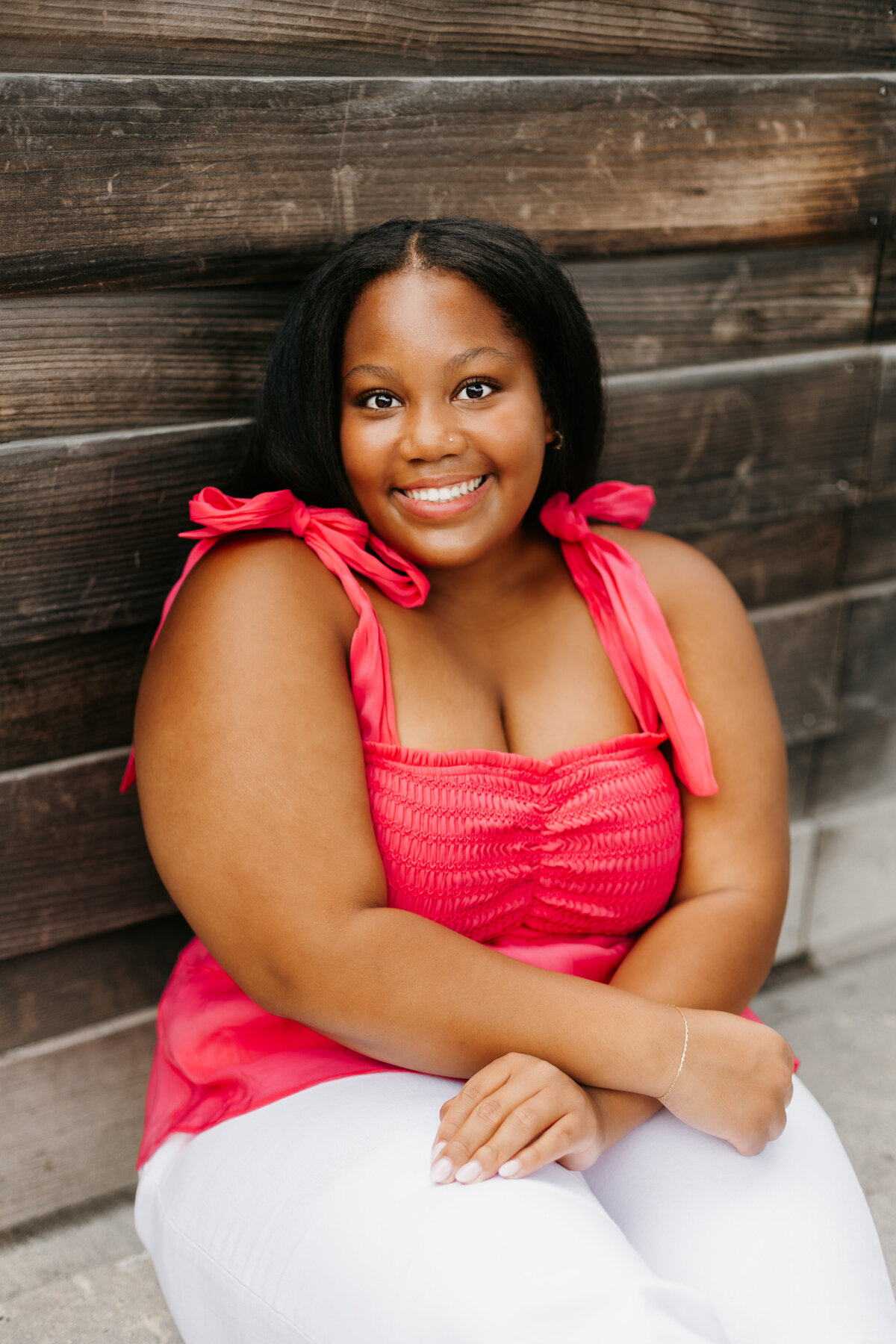 Senior girl in a pink top and white pants sitting in front of a wooden wall