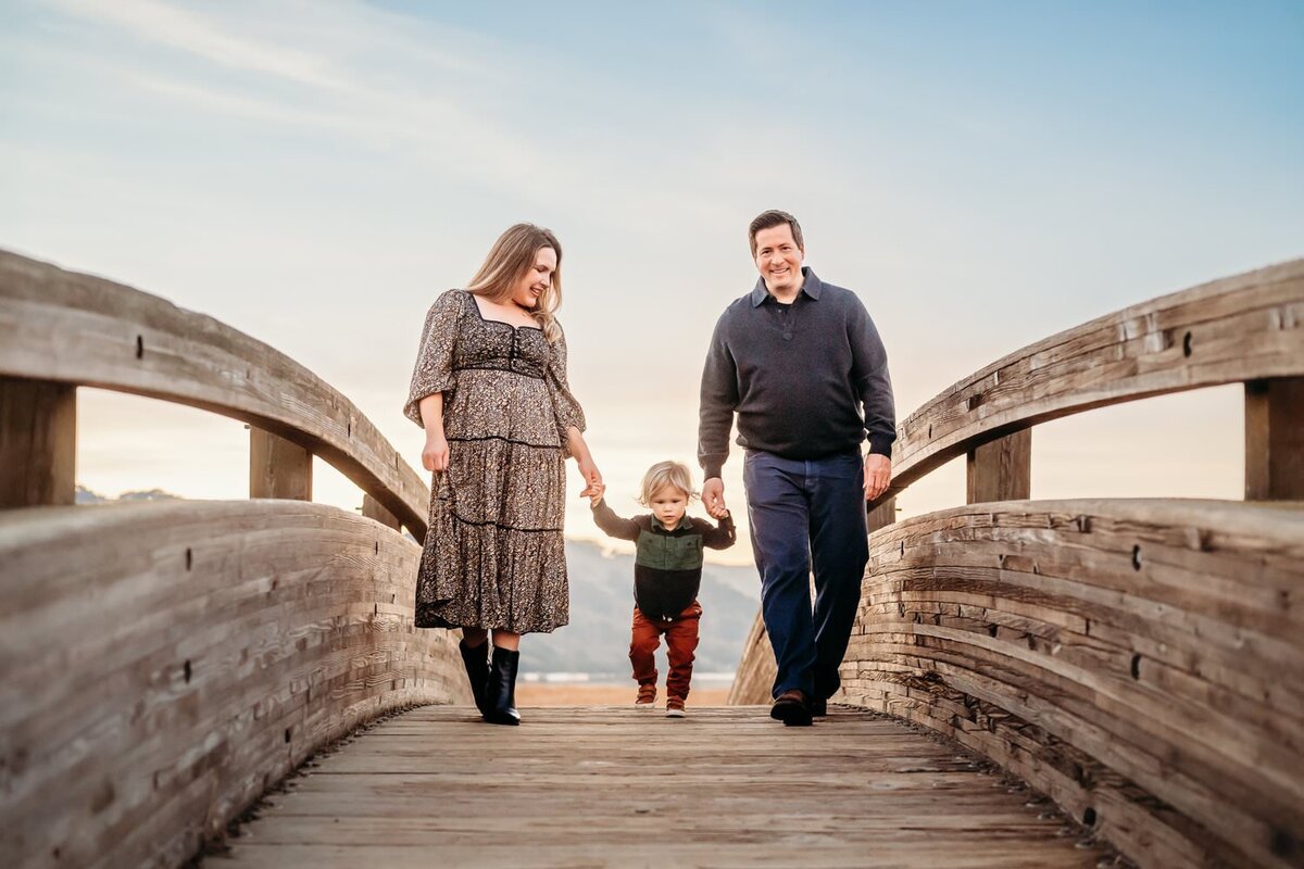 family of 3 walking over the scenic martinez waterfront bridge. there is a toddler holding both his parents hands as they walk towards the camera