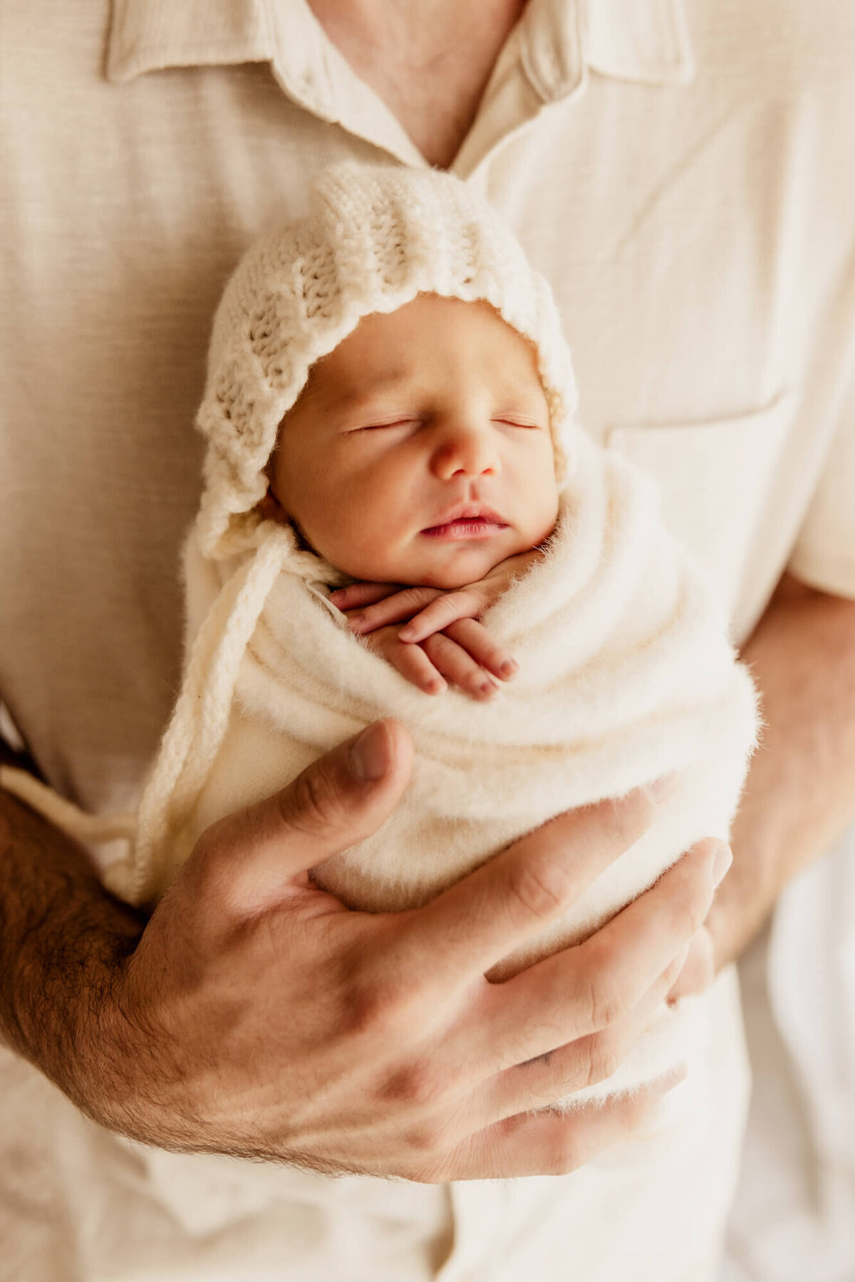 Baby girl with a white bonnet and white fuzzy swaddle held upright for a newborn photo in her father's arms.