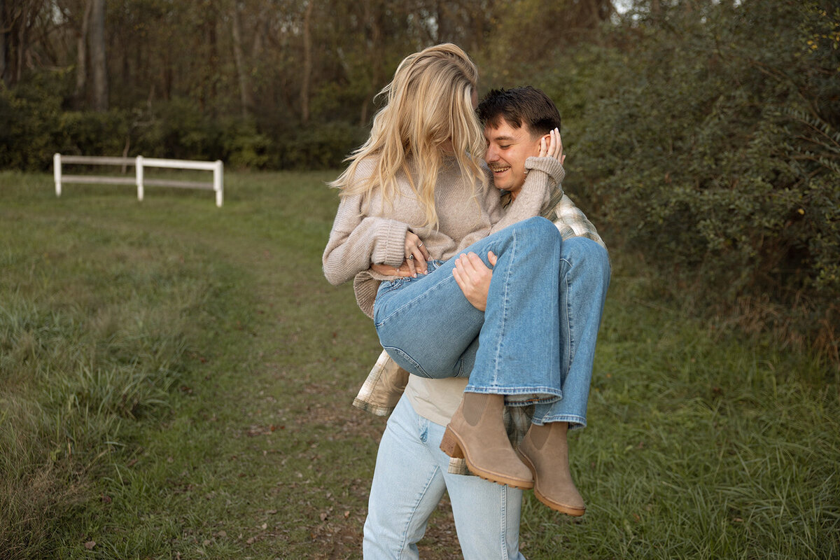 Man lifts up his fiance and twirls her around while they both laugh and smile at each other. 